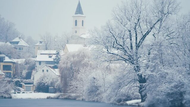A beautiful view of the Libocky rybnik and its' neighborhood in Prague. Snow-covered trees and houses on the banks of the small pond. A church is in the background. Slow-motion, pan-follow right.