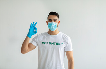 Fototapeta na wymiar Voluntary free work, charity grace concept. Male volunteer in medical mask and gloves showing OK gesture, light wall