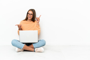 Young woman with a laptop sitting on the floor pointing finger to the laterals and happy