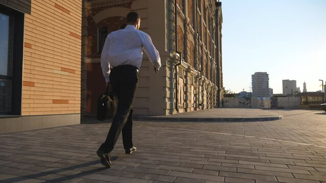 Successful manager in formal wear jogging on sidewalk. Young businessman with briefcase running along urban street. Office worker is late for meeting. Male entrepreneur hurries to work. Dolly shot