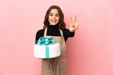 Little Pastry chef holding a big cake isolated on pink background smiling and showing victory sign