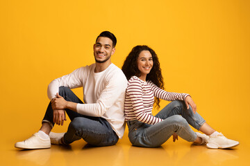 Cheerful Young Arab Couple Sitting Back To Back Over Yellow Studio Background