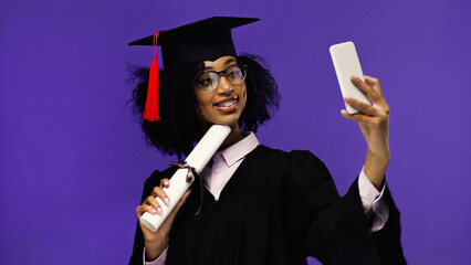 cheerful african american student with braces in graduation cap and gown taking selfie with rolled...