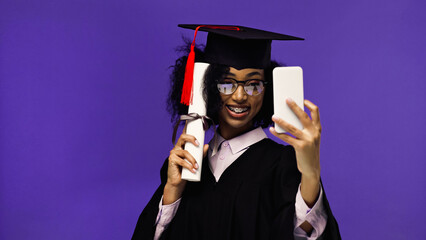 smiling african american student with braces in graduation cap and gown taking selfie with diploma isolated on purple.