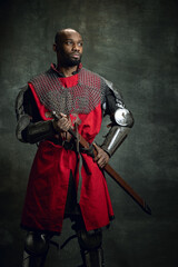 Fototapeta na wymiar Vintage style portrait of brutal dark skinned man, medieval warrior or knight with wounded face wearing armour isolated over dark background. Comparison of eras, history