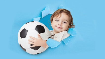 Cheerful cute european little kid with soccer ball looking through hole on blue paper, panorama