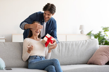 Smiling young european husband closes eyes to his wife and gives gift box in living room interior - obrazy, fototapety, plakaty