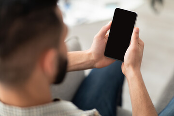 Unrecognizable millennial Arab guy using cellphone on couch at home, mockup for app or website design on screen