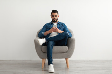 Young Arab guy using mobile phone, chatting online, sitting in armchair, surfing social media against white studio wall