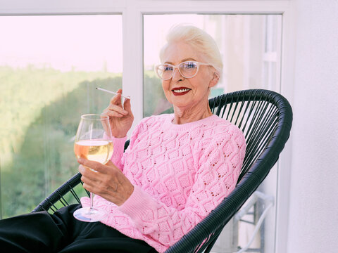 Old fashioned senior stylish woman sitting on terrace smoking cigarette with glass of white wine. Bad habit, addiction concept