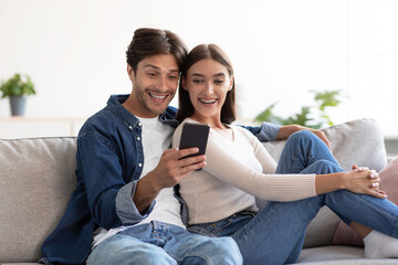 Smiling shocked young caucasian husband and wife look at smartphone, have meeting in living room