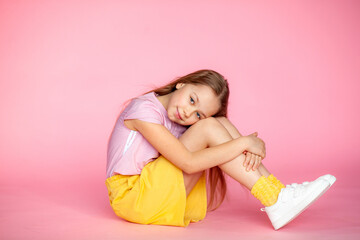 A funny little blonde girl of 10 years old in everyday bright clothes poses alone on a pink studio...