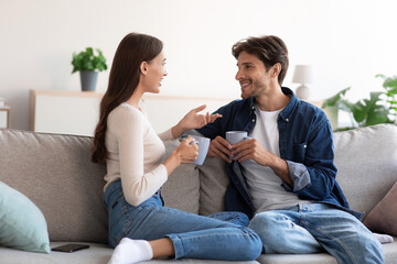 Smiling millennial caucasian male and female sit on sofa with cups of hot drink and talk in living room