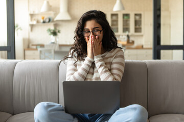 Young Caucasian woman looking at laptop screen, laughing, covering her mouth, watching funny movie on pc at home