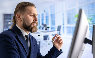 Bearded man in eyeglasses and formal suit working on modern pc while sitting at office desk. Caucasian mature worker looking at monitor with focused facial expression.