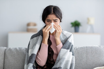 Unhappy sad young indian female in plaid suffering from fever and flu on sofa, blowing nose in...