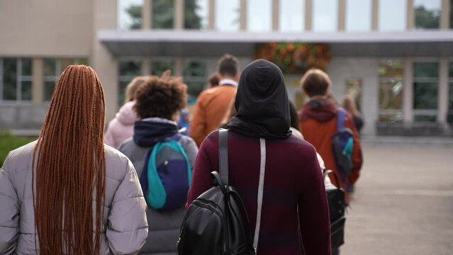Rear view of multi-ethnic secondary school pupils with backpacks walking to school building for lessons. Group of multiracial teenage schoolkids outdoor on the way to high school