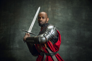 Obraz premium Vintage portrait of dark skinned medieval warrior wearing chain mail holding big sword isolated over dark vintage background. Comparison of eras, history. Side view