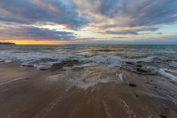 Golden hour sunset at Whitby beach. Warm sky and gentle, smooth waves along a sandy coastline with large rocks.