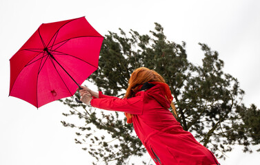 Eine Frau versucht bei starkem Wind ihren roten Regenschirm festzuhalten. Im Hintergrund steht ein...