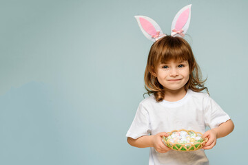 A cheerful girl with rabbit ears on her head with a basket of colored eggs in her hands on a blue background. Easter concept.