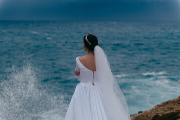 Fototapeta na wymiar Back view of brunette bride in white wedding dress and bridal veil on a cloudy day. Romantic beautiful bride posing near the sea with waves
