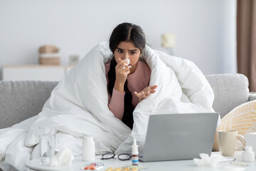 Unhappy young hindu woman wrapped in blanket, suffer from flu, on sofa, blow nose looks at computer
