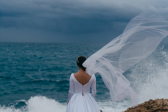 Back view of brunette bride in white wedding dress and bridal veil on a cloudy day. Romantic beautiful bride posing near the sea with waves