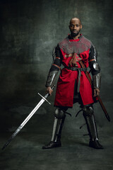 Vintage style portrait of brutal dark skinned man, medieval warrior or knight with wounded face wearing armour isolated over dark background. Comparison of eras, history