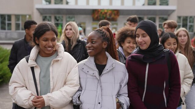 Group of happy high school students chatting while walking through school park after studies. Three multi-ethnic schoolgirls, indian, african american and arab female in hijab talking and smiling