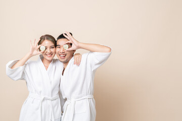 Happy Asian couple smile in bathrobe or spa suit with cucumber slice for facial mask together on brown isolated studio background.