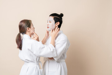 Happy Asian couple in bathrobe or spa suit doing sheet mask on face for facial treatment together...