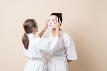 Happy Asian couple in bathrobe or spa suit doing sheet mask on face for facial treatment together on brown isolated studio background.