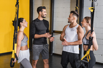 Smiling interracial people talking near suspension straps in gym.