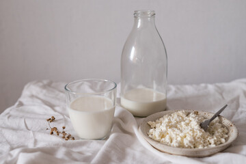 White still life of a bottle of milk, a glass of milk, a plate of cottage cheese. Items stand on a white tablecloth on a white background.