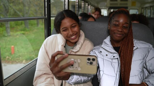 Cheerful mixed race and african american teenage girls taking selfie photo on smartphone while riding school bus after study. Joyful multi-ethnic classmates smiling while posing for portrait in bus