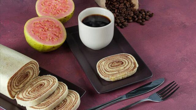 Slice of bolo de rolo surrounded by guava, cup and coffee beans on a red background.