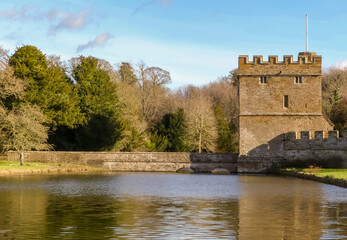 Public view of a medieval castellated gatehouse with bridge access over a moat. Water gently rippling. Woodland background under a blue and clouded sky. Landscape image with space for text. England. - 484699610