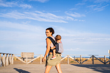 Mother carrying a baby on her back in a baby carrier next to the beach