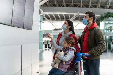 Fototapeta na wymiar Side view of the parents wearing protective masks standing in front of the timetable board and waiting for the flight at the airport while their daughter pointing with finger. 