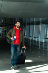Fototapeta na wymiar Full length view of the confident young man is going through the hall with suitcase and waiting for the flight in airport. Traveling concept