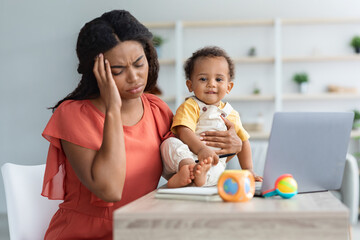 Tired Black Mother With Baby Suffering Headache While Using Laptop At Home