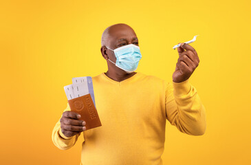 Portrait of senior black male tourist in face mask holding passport, tickets and toy plane on orange studio background