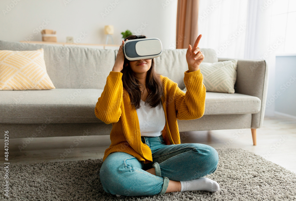 Wall mural Joyful arab young woman wearing modern VR glasses while sitting on floor in living room, pointing at copy space