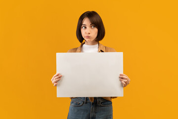 Fototapeta na wymiar Making decision. Doubtful young lady holding empty blank board, posing on yellow studio background