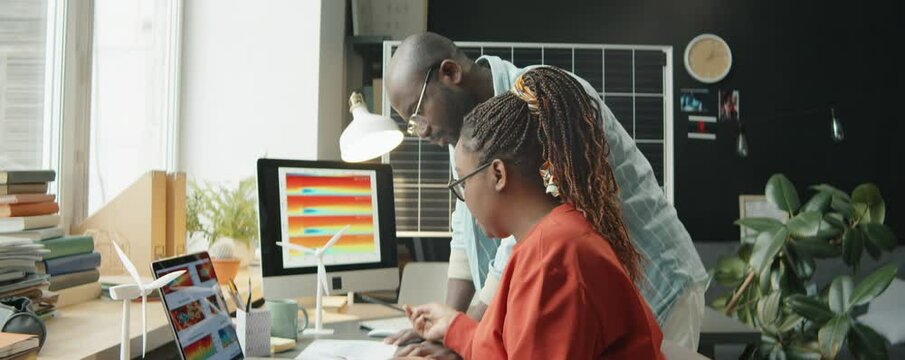 African American female engineer and her male colleague examining papers and discussing slide on computer while working together on renewable energy project