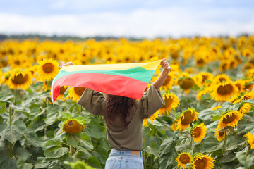 Woman holding flag of Lithuania in a sunflowers field. Back view. Lithuanian Flag Day. Independence restoration Day. Travel and love Lithuania concept. Selective focus. - obrazy, fototapety, plakaty