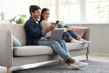 Glad cheerful millennial european man and woman sitting on sofa with laptop and looking at smartphone