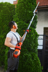 Muscular caucasian gardener trimming overgrown thujas with electric cutter outdoors. Man in working...