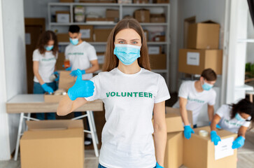 Female activist wearing medical mask, working at volunteer center, showing thumb up at camera